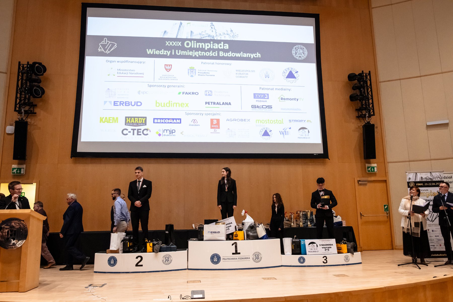 Ceremonia wręczenia nagród podczas finału XXXIX Olimpiady Wiedzy i Umiejętności Budowlanych - laureaci stoją na podium na scenie, a w tle widoczny jest ekran z nazwą wydarzenia i logotypami sponsorów.