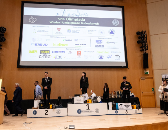 Ceremonia wręczenia nagród podczas finału XXXIX Olimpiady Wiedzy i Umiejętności Budowlanych - laureaci stoją na podium na scenie, a w tle widoczny jest ekran z nazwą wydarzenia i logotypami sponsorów.