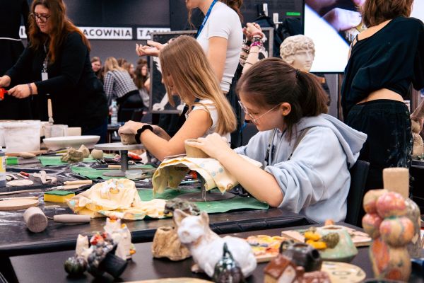 Arena Zawodów, czyli dlaczego warto mieć w ręku konkretny fach? Grupa młodych osób pracuje przy stole z ceramiką, formując i dekorując gliniane figurki w kreatywnej atmosferze warsztatów.