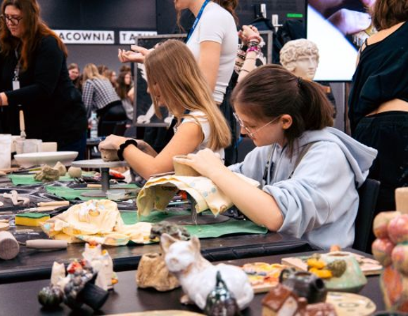 Arena Zawodów, czyli dlaczego warto mieć w ręku konkretny fach? Grupa młodych osób pracuje przy stole z ceramiką, formując i dekorując gliniane figurki w kreatywnej atmosferze warsztatów.