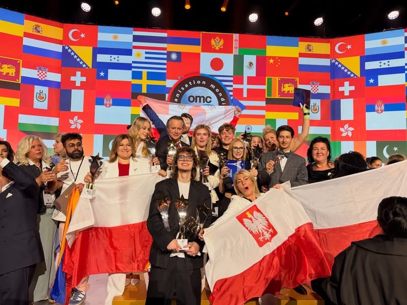 Grupa uczestników z biało-czerwonymi flagami Polski pozuje do wspólnego zdjęcia na tle flag państw świata podczas międzynarodowego wydarzenia.