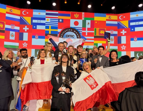 Grupa uczestników z biało-czerwonymi flagami Polski pozuje do wspólnego zdjęcia na tle flag państw świata podczas międzynarodowego wydarzenia.
