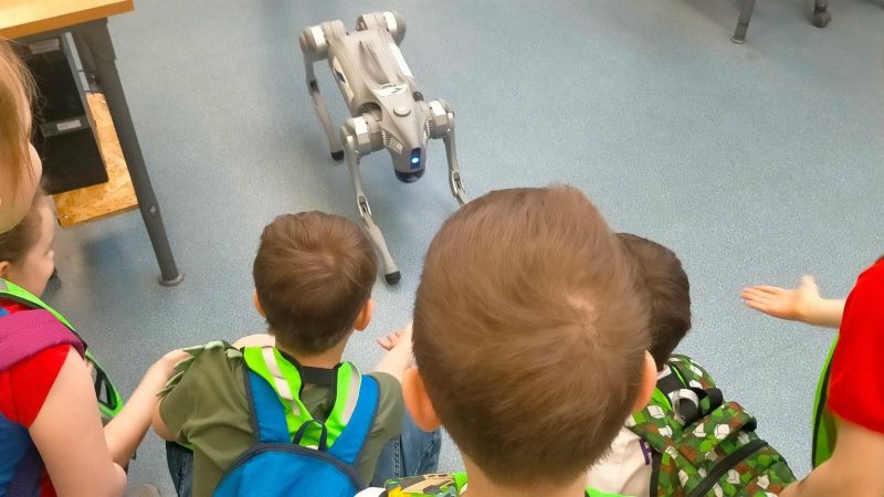 "Przygodę z zawodem czas zacząć" Kilkoro dzieci siedzi na podłodze i obserwuje czworonożnego robota przypominającego psa w szkolnej sali.