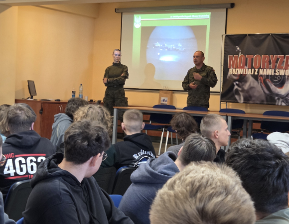 Na grafice znajdują się uczniowie siedzący w klasie i oglądający prezentację multimedialną