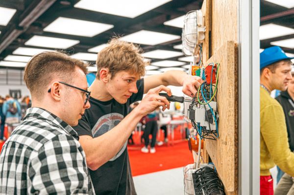 Arena Zawodów - warto postawić na naukę konkretnego fachu Na zdjęciu widać dwóch młodych mężczyzn pracujących przy panelu z instalacją elektryczną. Jeden z nich używa śrubokręta, koncentrując się na podłączeniu przewodów, podczas gdy drugi uważnie obserwuje. W tle znajdują się inne osoby, co sugeruje, że scena ma miejsce na targach.