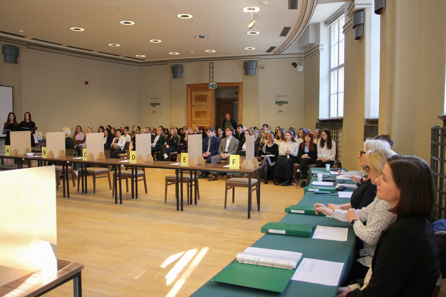 Na zdjęciu znajduje się komisja zasiadająca za stołem oraz uczniowie oczekujący na rozpoczęcie konkursu.