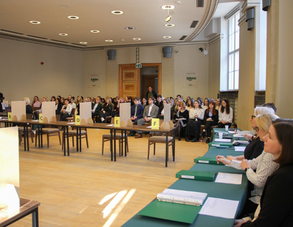 Na zdjęciu znajduje się komisja zasiadająca za stołem oraz uczniowie oczekujący na rozpoczęcie konkursu.