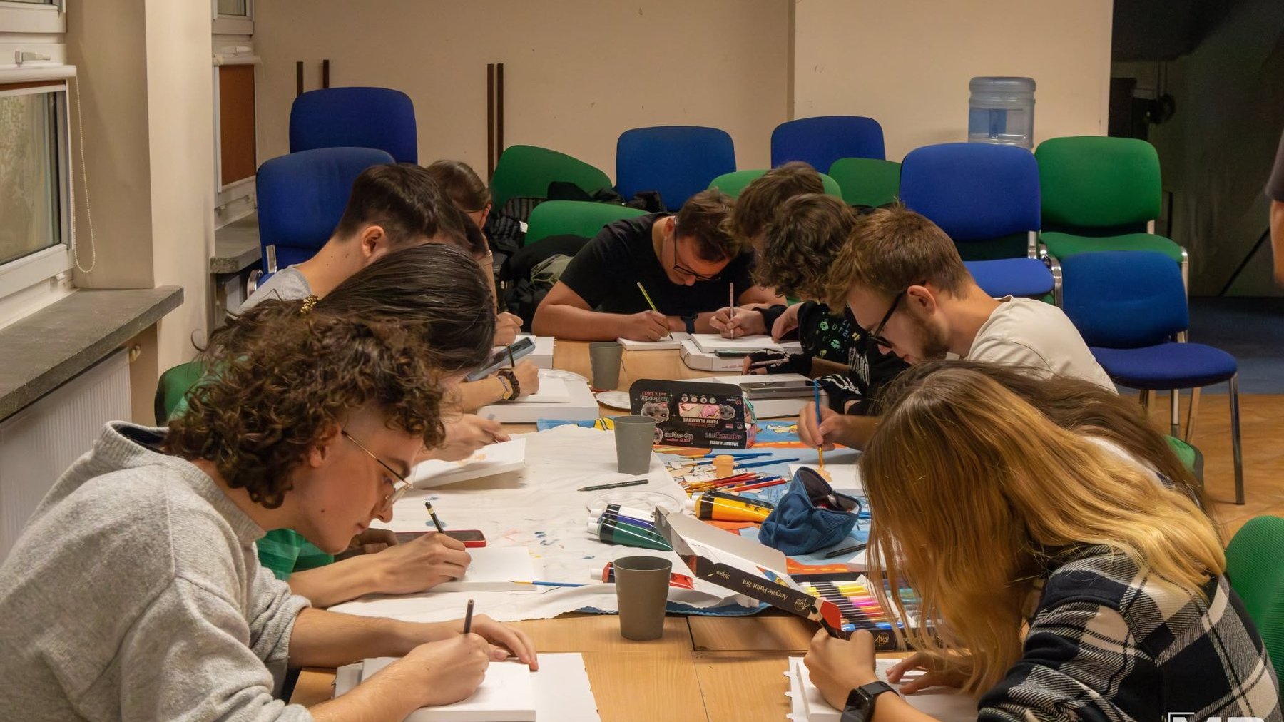 Na grafice znajduje się młodzież, siedzą w klasie szkolnej i rysują