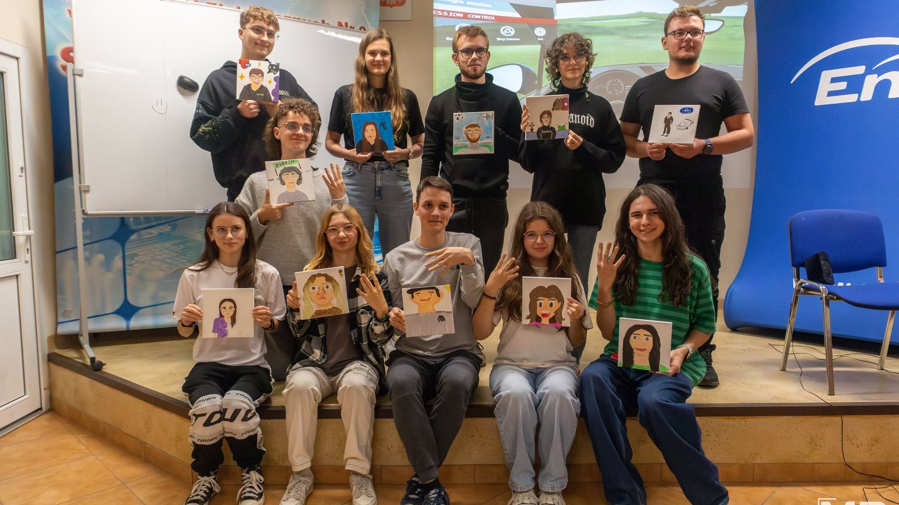 Na grafice znajduje się młodzież, siedzą w klasie szkolnej i pokazują swoje prace, namalowane podobizny