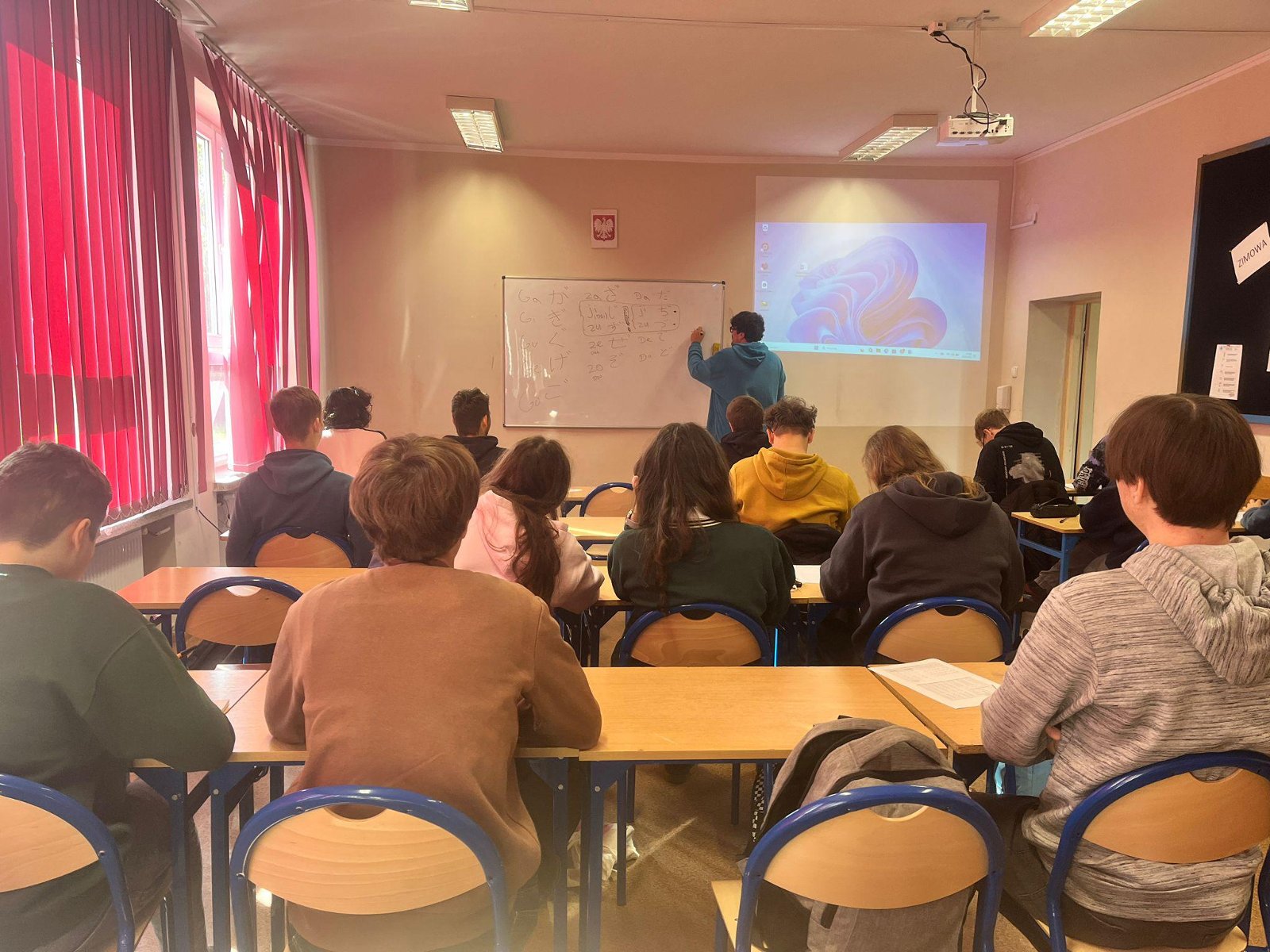 Nauczyciel pisze na tablicy znaki japońskiego alfabetu podczas lekcji. Uczniowie siedzą w ławkach i śledzą przebieg zajęć. - grafika artykułu