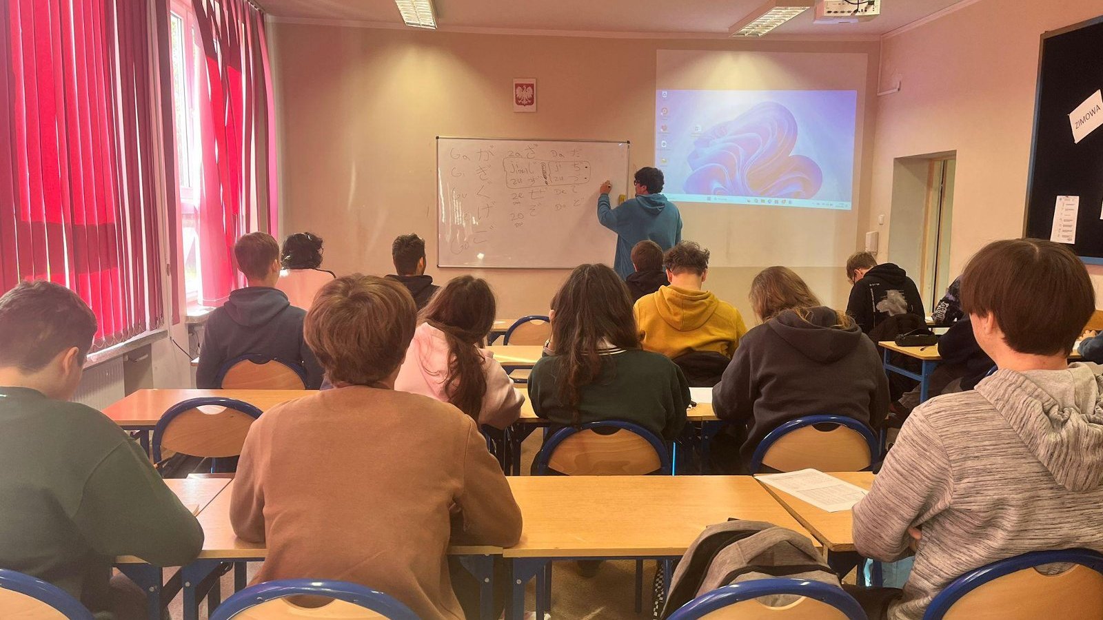 Nauczyciel pisze na tablicy znaki japońskiego alfabetu podczas lekcji. Uczniowie siedzą w ławkach i śledzą przebieg zajęć.