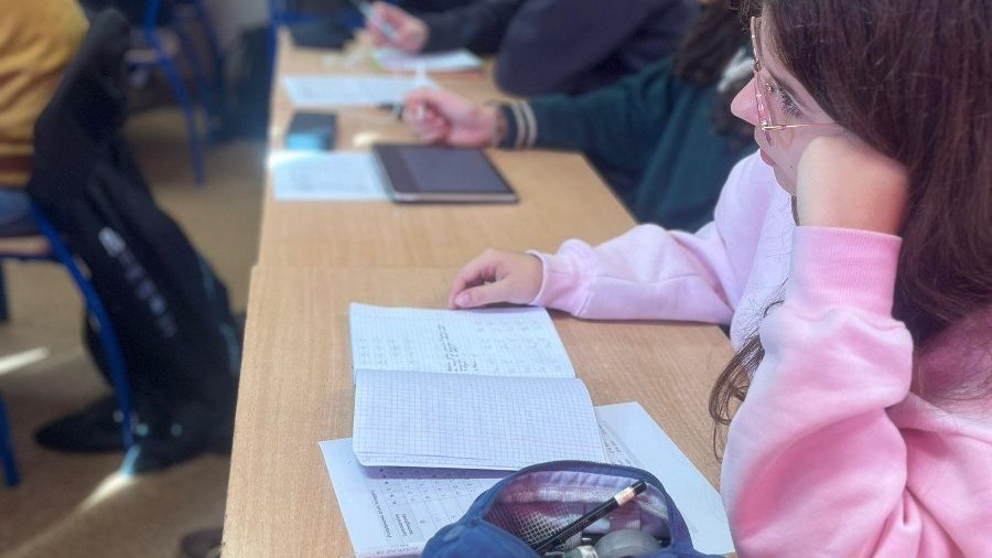 Uczniowie siedzą w ławkach i uważnie notują podczas zajęć z języka japońskiego. Na pierwszym planie widać otwarty zeszyt i piórnik.