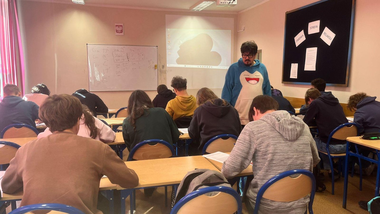 Prowadzący zajęcia języka japońskiego pomaga uczniom w klasie. Na tablicy widoczne są japońskie znaki, a uczniowie pracują nad zadaniami.