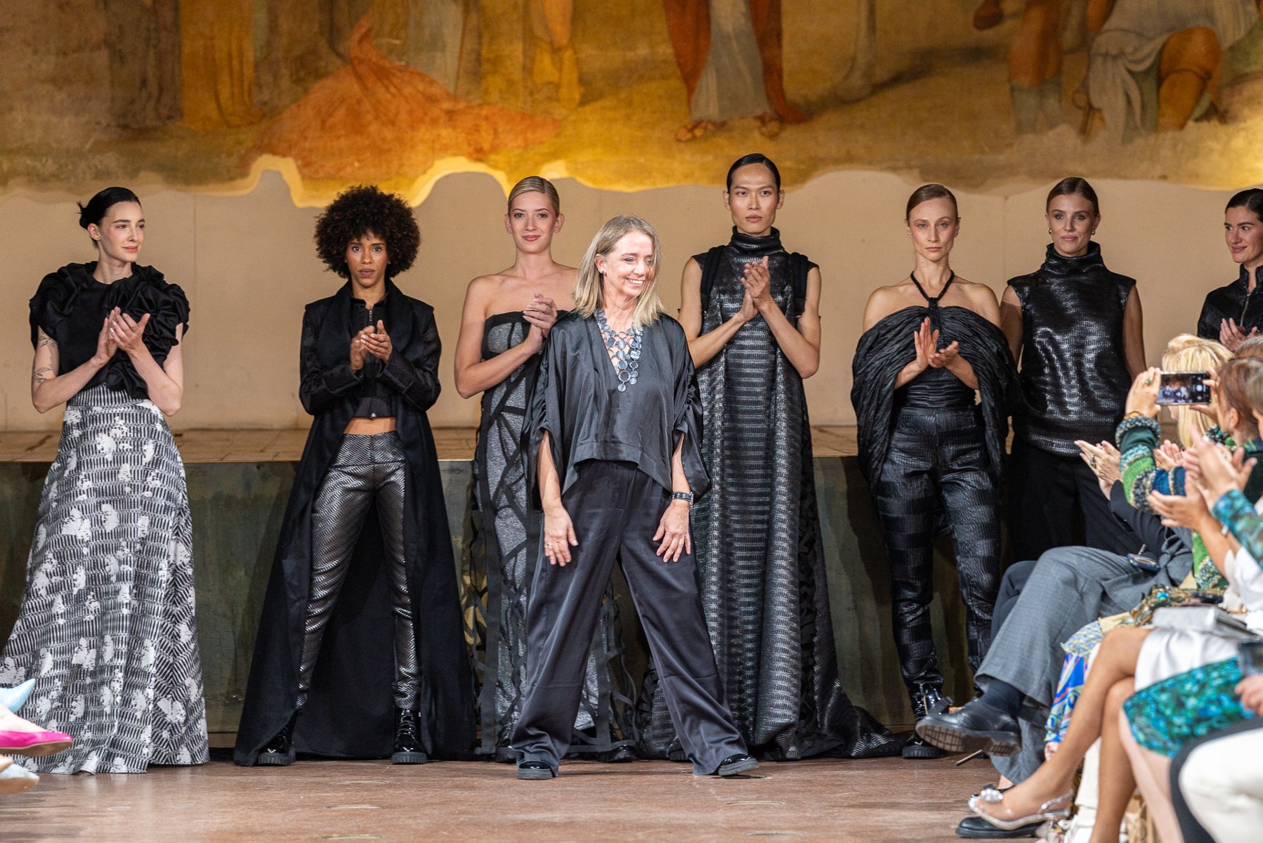 Projektantka stoi na wybiegu wśród modelek w czarnych kreacjach, odbierając brawa po pokazie mody. - grafika artykułu