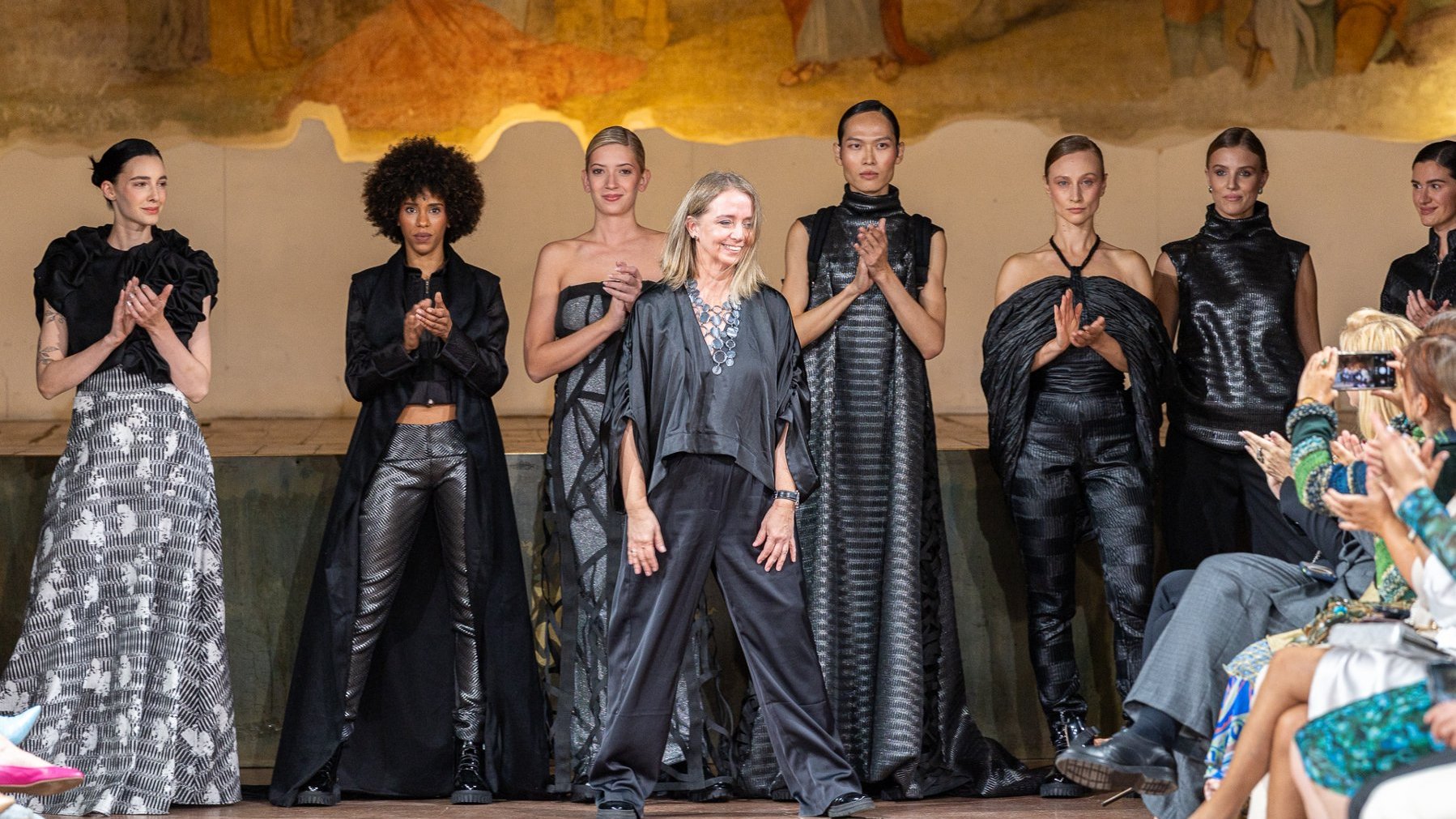 Projektantka stoi na wybiegu wśród modelek w czarnych kreacjach, odbierając brawa po pokazie mody.