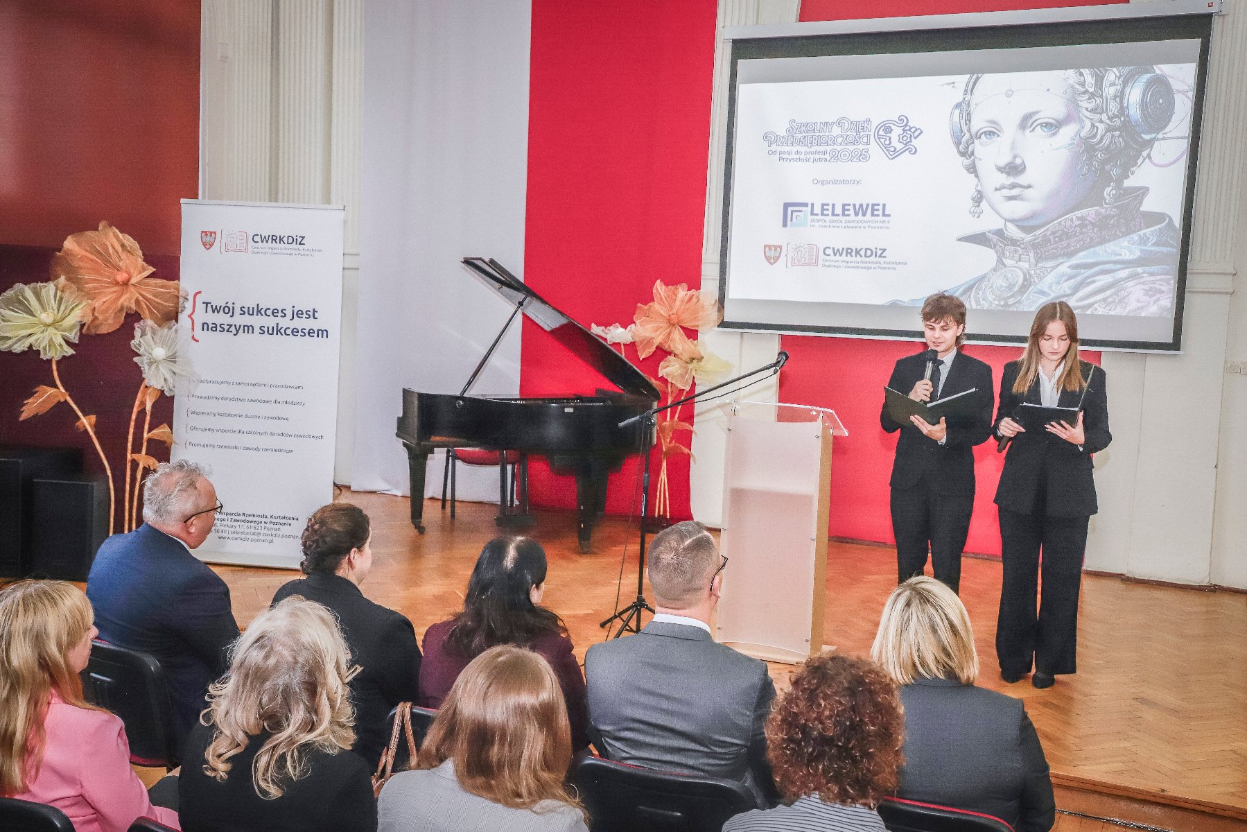 Aula szkolna, widoczni są goście audytorium oraz dwóch prowadzących uczniów. - grafika artykułu