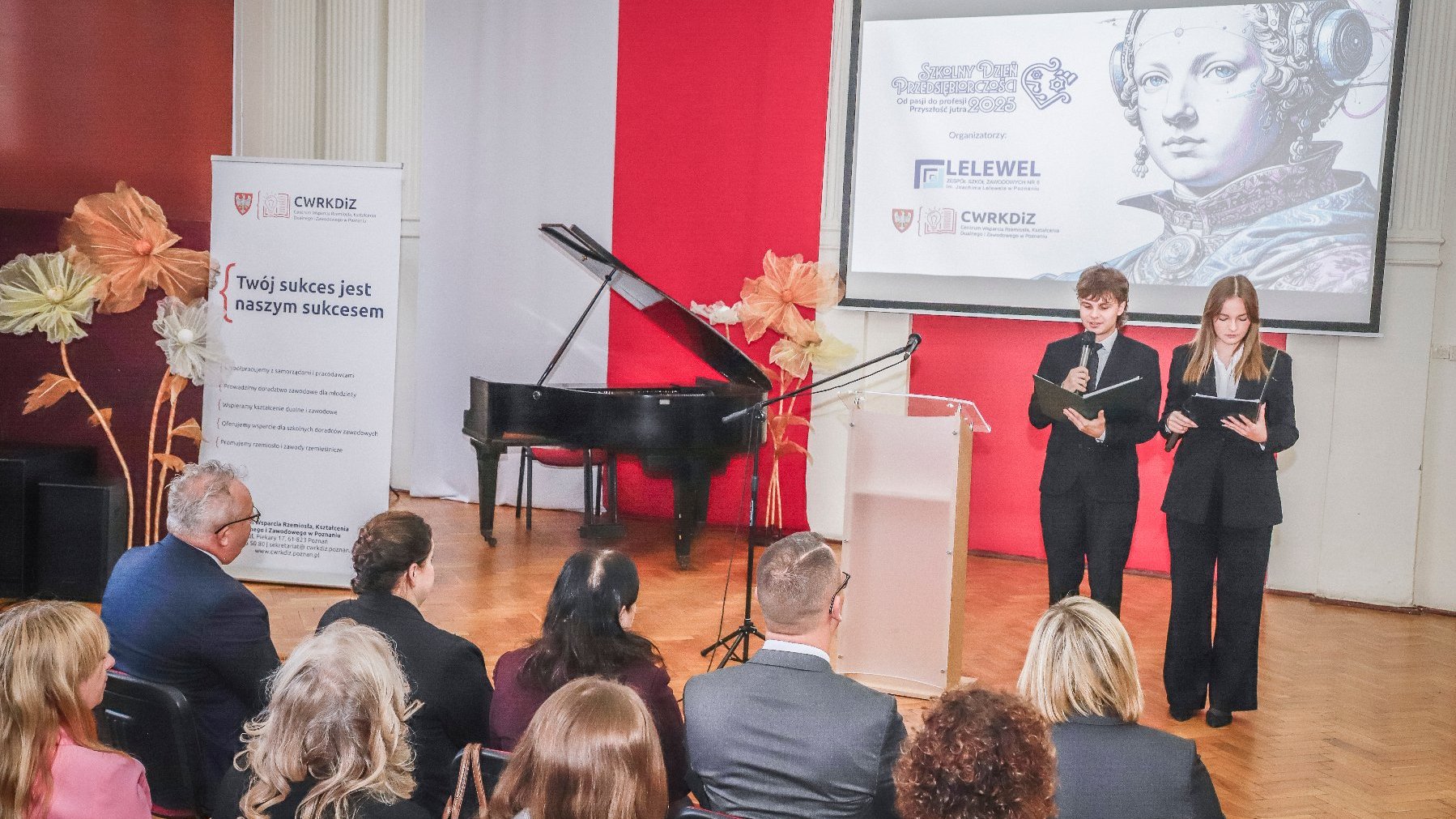 Aula szkolna, widoczni są goście audytorium oraz dwóch prowadzących uczniów.