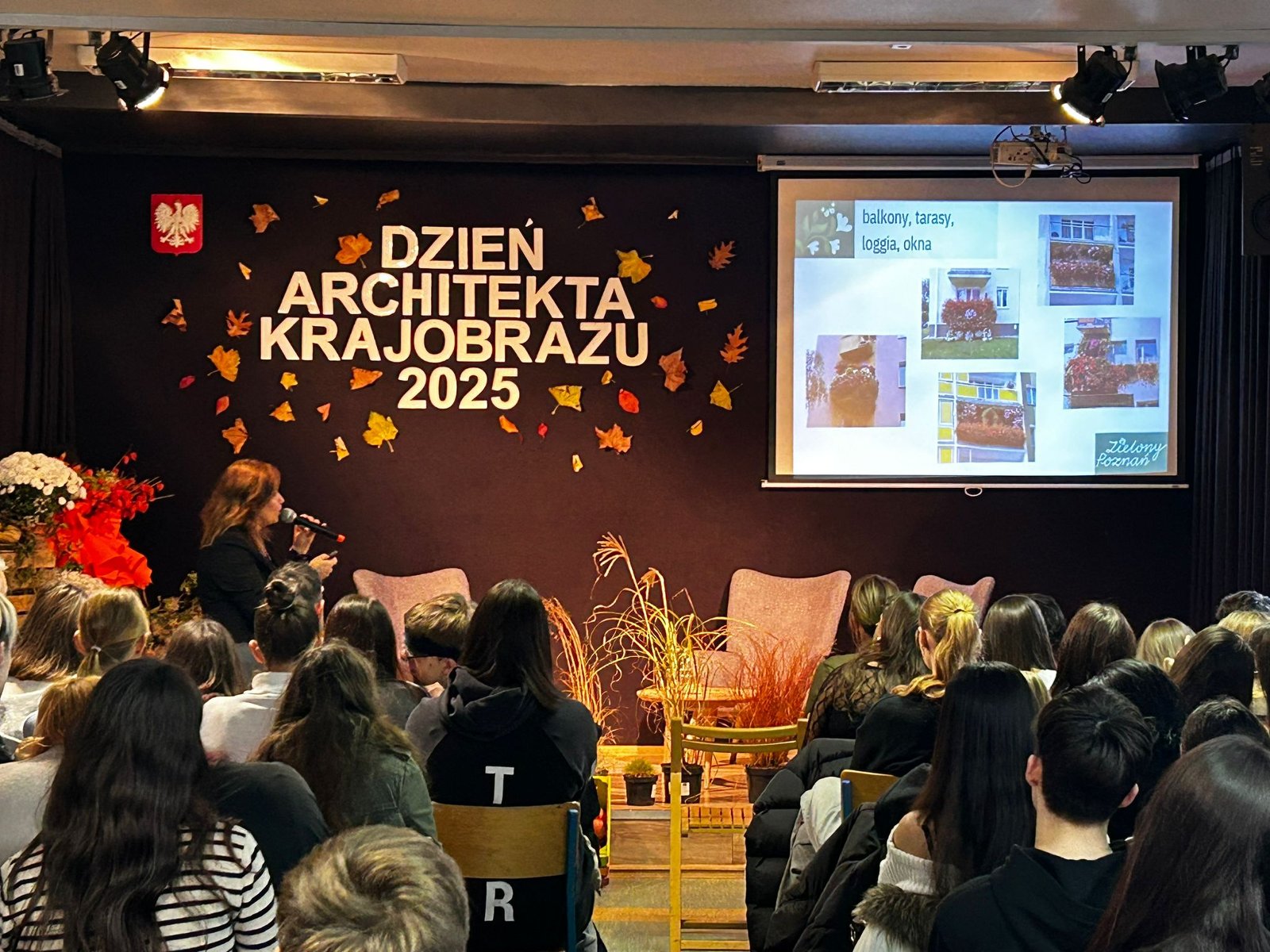 Na zdjęciu przedstawiono aulę szkolną, na widowni siedzą uczniowie i oglądają prezentację multimedialną ze zdjęciami - grafika artykułu