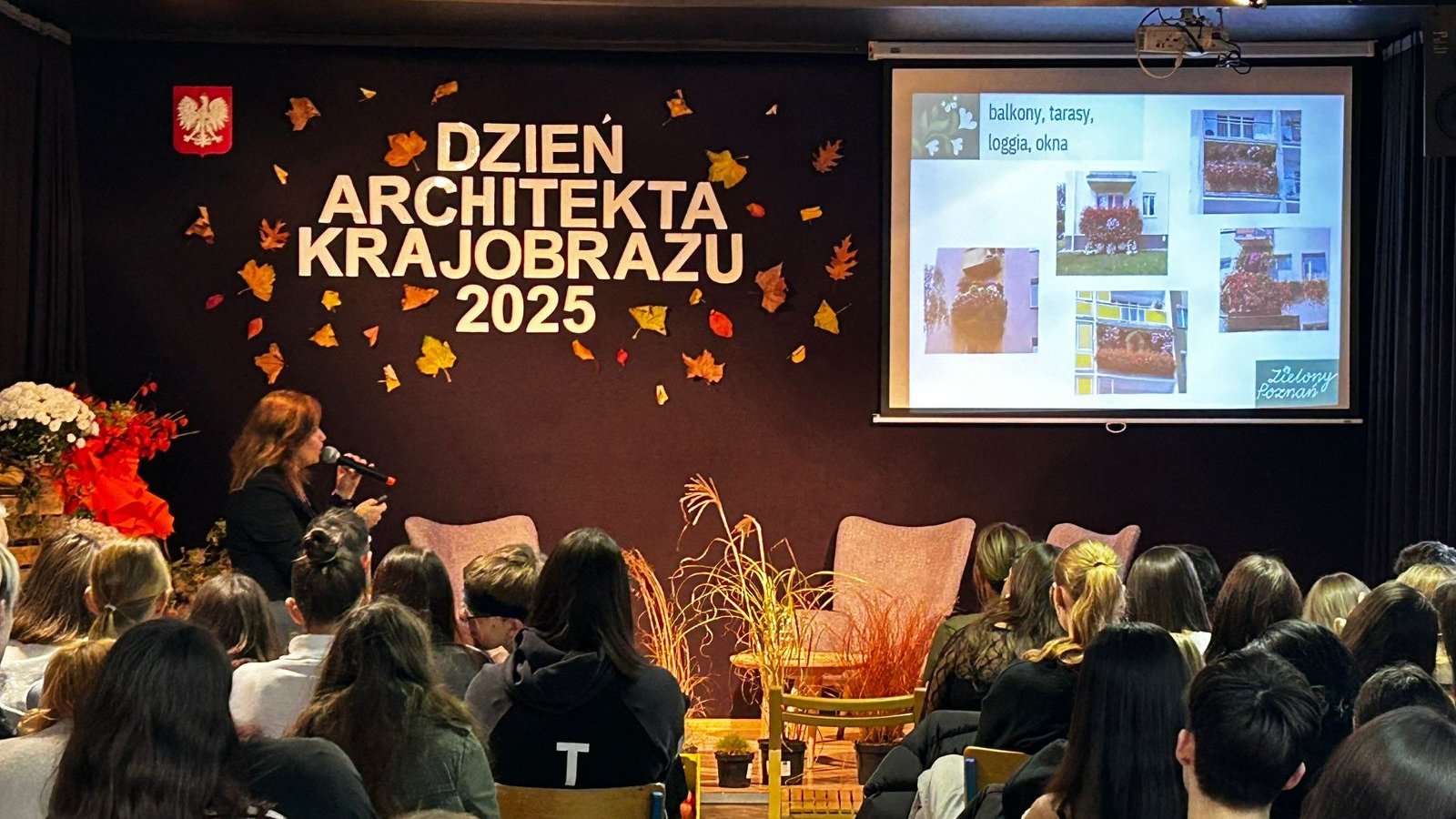 Na zdjęciu przedstawiono aulę szkolną, na widowni siedzą uczniowie i oglądają prezentację multimedialną ze zdjęciami