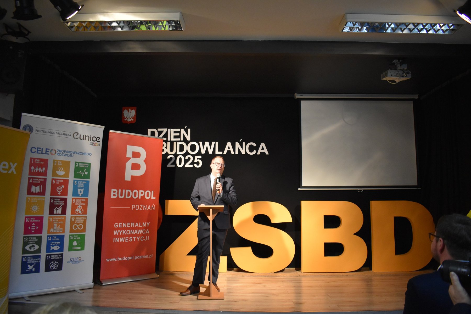 Na czarnym tle sceny przemawia Dyrektor Zespołu Szkół Budowlano-Drzewnych - grafika artykułu