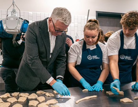 Prezydent Jacek Jaśkowiak wraz z młodzieżą w fartuchach, formuje ciasto w kuchni, uczestnicząc w warsztatach kulinarnych w Branżowym centrum.