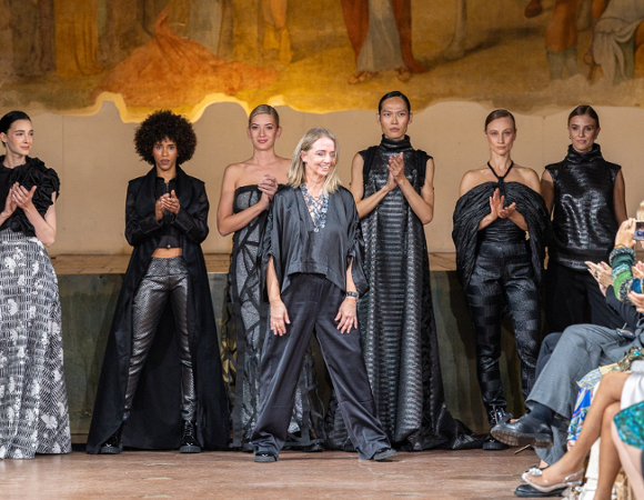 Tydzień mody w Mediolanie. Projektantka stoi na wybiegu wśród modelek w czarnych kreacjach, odbierając brawa po pokazie mody.