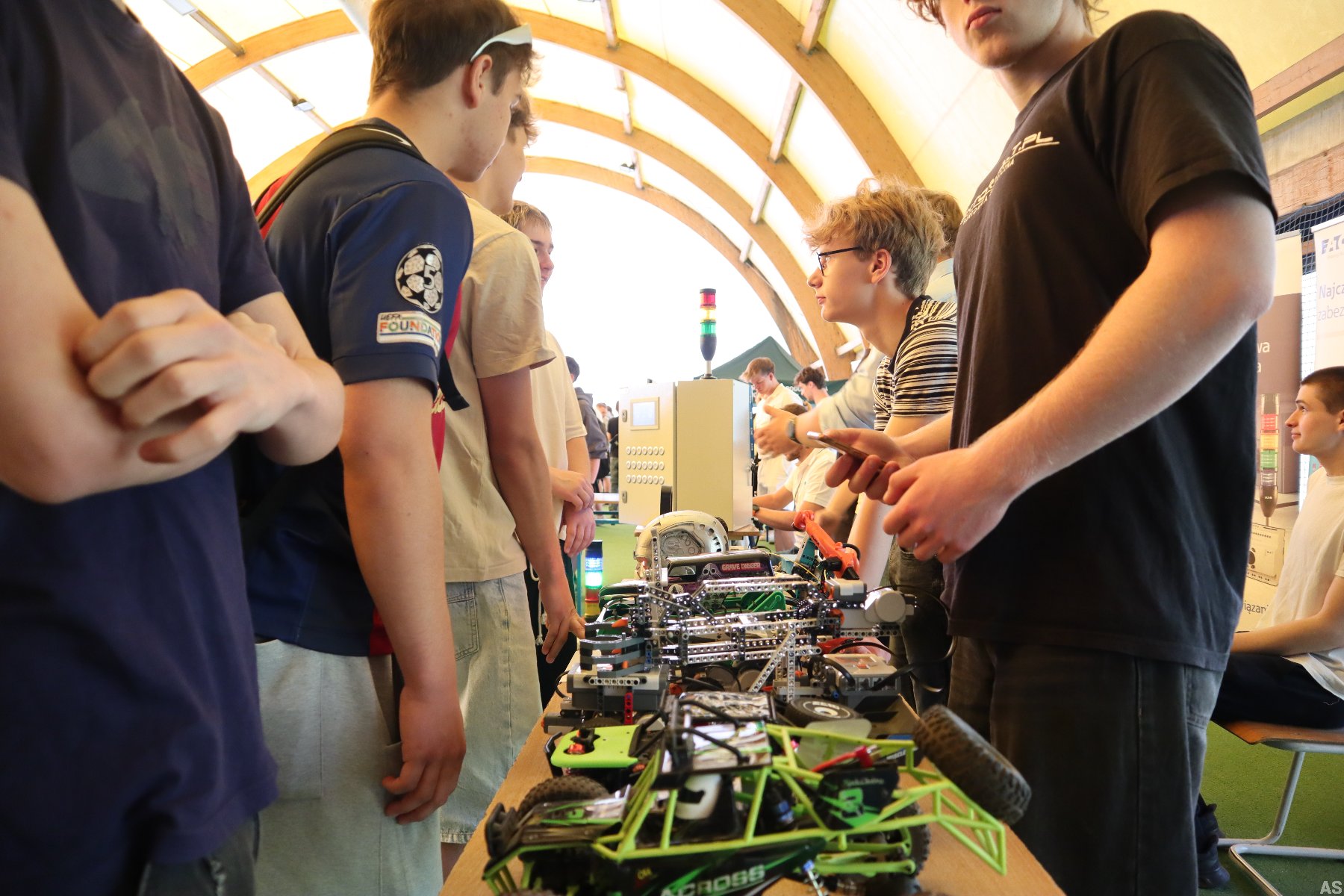 Uczniowie prezentują projekty techniczne i modele robotyczne podczas szkolnego wydarzenia. Na pierwszym planie widać zdalnie sterowany pojazd i konstrukcje mechaniczne ustawione na stole.