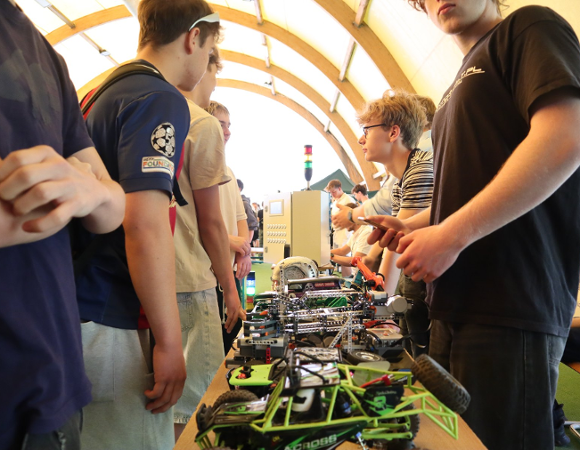 Uczniowie prezentują projekty techniczne i modele robotyczne podczas szkolnego wydarzenia. Na pierwszym planie widać zdalnie sterowany pojazd i konstrukcje mechaniczne ustawione na stole.