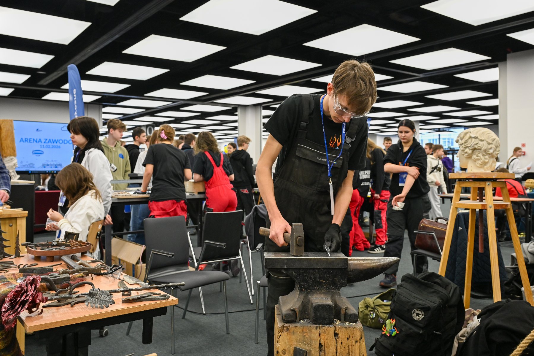 Zdjęcie przedstawia dwie młode dziewczyny ubierające manekina. W tle widać innych młodych ludzi.